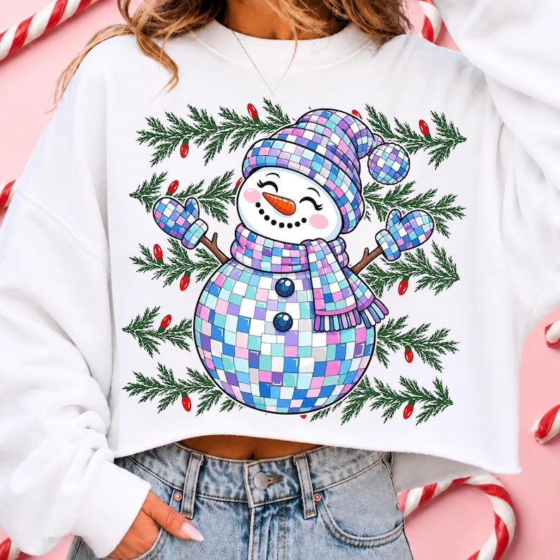 May include: A white long-sleeve sweatshirt featuring a graphic of a snowman made of iridescent disco ball tiles. The snowman wears a matching checkered hat, scarf, and mittens, set against a background of green pine branches and red light bulbs.