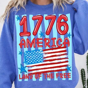 Może przedstawiać: Niebieska bluza z okrągłym dekoltem z nadrukiem graficznym zawierającym czerwony napis 1776, słowo AMERICA oraz frazę LAND OF THE FREE. Wz&oacute;r zawiera stylizowaną flagę amerykańską, jasnoniebieskie pionowe paski oraz kilka niebieskich gwiazdek.