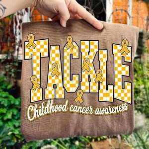 May include: A brown t-shirt featuring the word TACKLE in large, yellow and white checkered block letters. Several yellow awareness ribbons are integrated into the design. Below the word, the text Childhood cancer awareness is printed in white.