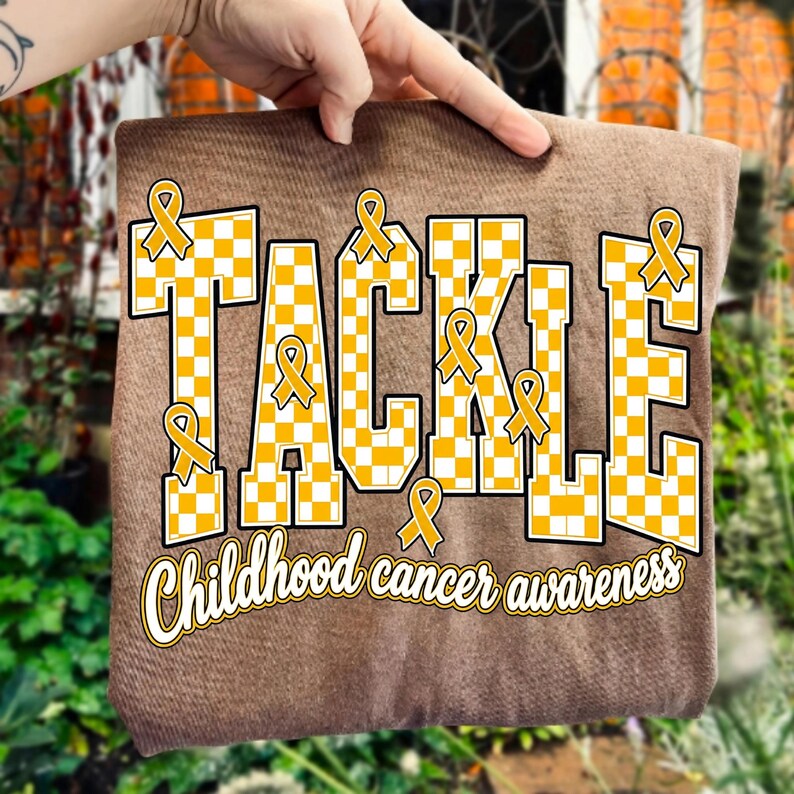 May include: A brown t-shirt featuring the word TACKLE in large, yellow and white checkered block letters. Several yellow awareness ribbons are integrated into the design. Below the word, the text Childhood cancer awareness is printed in white.