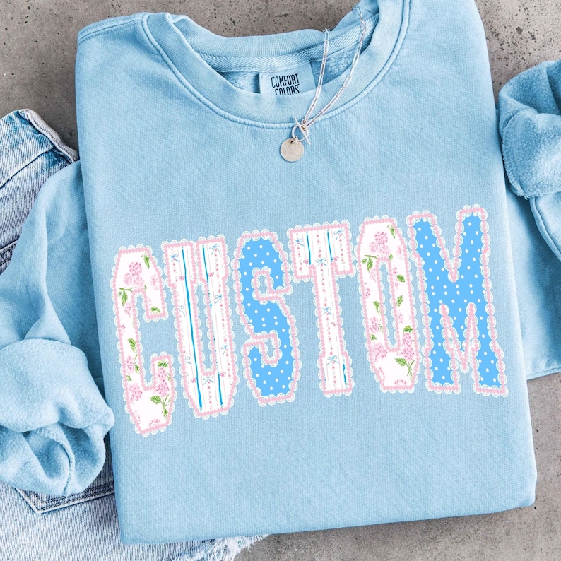 May include: A light blue sweatshirt with the word "CUSTOM" in large, decorative letters. The letters have a scalloped pink border and are filled with floral and polka dot patterns. A silver necklace with a small pendant is visible.