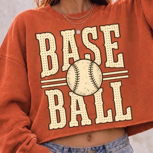 Puede incluir: Una sudadera corta de color naranja quemado con un gran estampado gr&aacute;fico que muestra el texto BASE BALL en una fuente serif de color crema con puntos. Una ilustraci&oacute;n central de una pelota de b&eacute;isbol con costuras visibles separa las palabras.