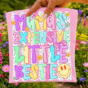 May include: A light pink rectangular fabric item featuring a black and white polka dot striped background. The text reads MAMA'S EXPENSIVE LITTLE BESTIE in colorful, checkered, and polka-dotted letters, accented with small flowers, bows, and a smiley face.