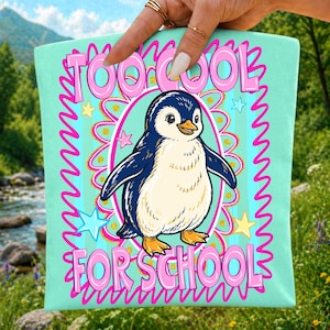 Pu&ograve; includere: Una borsa tote verde menta chiaro con un'illustrazione centrale di un pinguino all'interno di una cornice ovale rosa. Il testo "TOO COOL FOR SCHOOL" &egrave; stampato in grandi lettere rosa sopra e sotto il pinguino, circondato da piccole stelle gialle e blu.