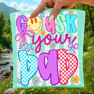 Op de afbeelding: Een lichtgroen t-shirt met een kleurrijke afbeelding en de tekst "GO ASK YOUR DAD". Het ontwerp bevat een geel smileygezicht, roze en blauw geblokte letters, kleine bloemen, sterren en roze strikjes op een witte achtergrond met zwarte stippen.