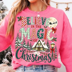 May include: A bright pink crewneck sweatshirt featuring a festive graphic with the text BELIEVE IN THE MAGIC OF CHRISTMAS. The design includes a snowy cabin, a street lamp, pine trees, holly, snowflakes, and a silhouette of Santa in a sleigh.