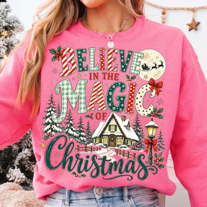 May include: A bright pink crewneck sweatshirt featuring a festive graphic with the text BELIEVE IN THE MAGIC OF CHRISTMAS. The design includes a snowy cabin, a street lamp, pine trees, holly, snowflakes, and a silhouette of Santa in a sleigh.