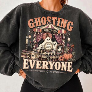 Pode incluir: Uma camisola cinzento-carv&atilde;o de gola redonda com uma impress&atilde;o gr&aacute;fica de um fantasma de turbante atr&aacute;s de uma bola de cristal, um gato preto, cartas de tarot e uma ab&oacute;bora. O texto diz GHOSTING EVERYONE, SPIRIT LINE, HELLO? YOU THERE? ANSWER ME e NO APPOINTMENTS NO APPARITIONS.