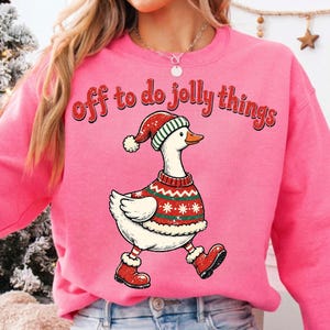 May include: A bright pink crewneck sweatshirt featuring a graphic of a white goose wearing a red and green patterned holiday sweater, a matching Santa hat, and red boots. The text "off to do jolly things" is printed in red above the goose.