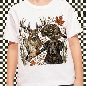 May include: A white t-shirt featuring a graphic print of a deer, a flying mallard duck, a camouflage baseball cap, and a black Labrador retriever wearing a brown collar, all surrounded by cattails and autumn leaves.