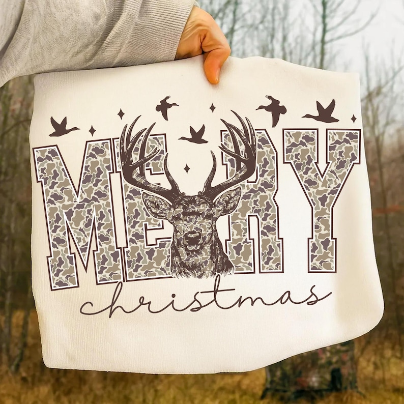 May include: Cream-colored tote bag featuring the words "MERRY Christmas" with a deer graphic. The letters of "MERRY" have a camouflage pattern. The deer has large antlers and is centered above the word "Christmas". Decorative birds and stars are above the word "MERRY".