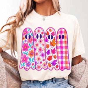 May include: A cream-colored t-shirt featuring a graphic of four ghosts standing side-by-side. Each ghost has a unique pattern: the first has large pink flowers, the second has small pink daisies, the third has autumn leaves, and the fourth has a pink plaid pattern.