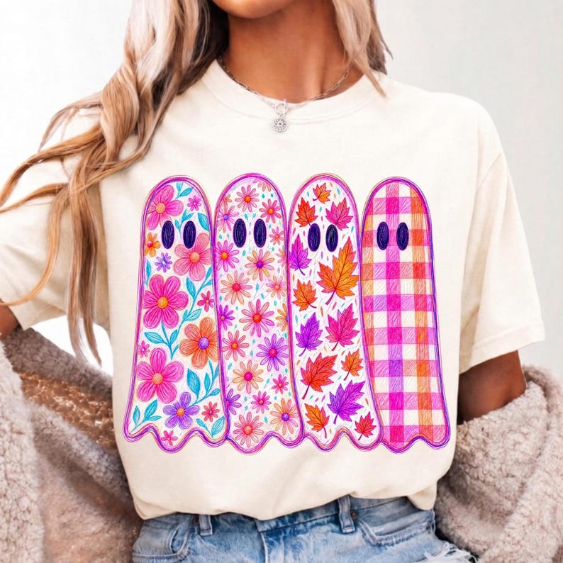 May include: A cream-colored t-shirt featuring a graphic of four ghosts standing side-by-side. Each ghost has a unique pattern: the first has large pink flowers, the second has small pink daisies, the third has autumn leaves, and the fourth has a pink plaid pattern.