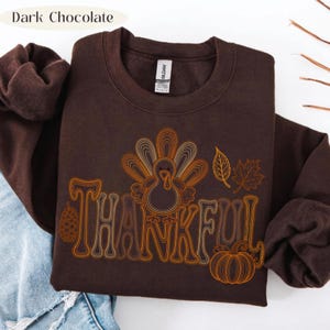 May include: A dark chocolate brown crewneck sweatshirt featuring a graphic print with the word THANKFUL in a stylized font. The design includes a turkey, a pinecone, a pumpkin, and autumn leaves in shades of orange, yellow, and cream.