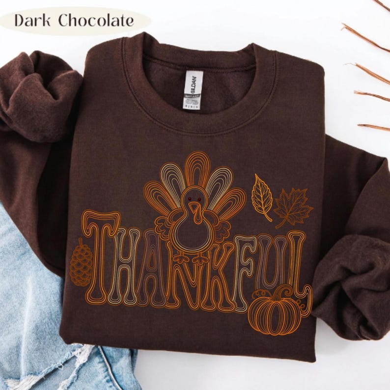 May include: A dark chocolate brown crewneck sweatshirt featuring a graphic print with the word THANKFUL in a stylized font. The design includes a turkey, a pinecone, a pumpkin, and autumn leaves in shades of orange, yellow, and cream.