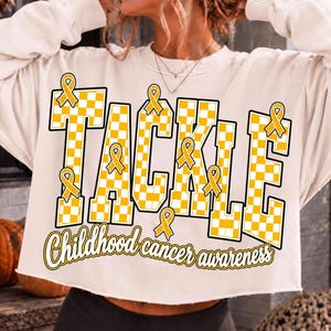 May include: A cream-colored long-sleeve sweatshirt with a raw hem featuring the word TACKLE in a yellow and white checkered pattern. The design includes yellow awareness ribbons and the text Childhood cancer awareness in yellow script.