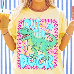 May include: A pale yellow t-shirt featuring a graphic of a green Tyrannosaurus Rex in the centre. The text "OUT THE DOOR" is printed in large, light blue letters around the dinosaur. The design includes pink squiggly borders and stars.