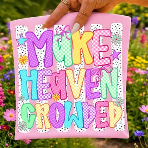 May include: A pink tote bag featuring the text MAKE HEAVEN CROWDED in colorful, patterned block letters. The design includes polka dot stripes, small flowers, stars, and bows in shades of purple, yellow, teal, and pink against a white background.