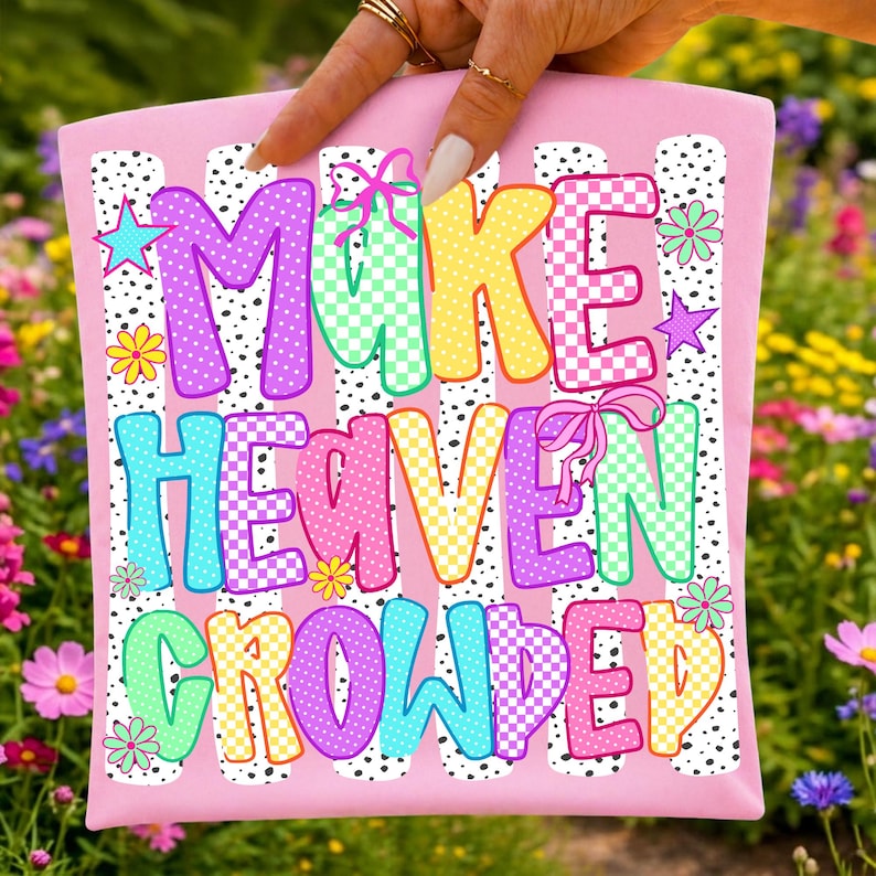 May include: A pink tote bag featuring the text MAKE HEAVEN CROWDED in colorful, patterned block letters. The design includes polka dot stripes, small flowers, stars, and bows in shades of purple, yellow, teal, and pink against a white background.