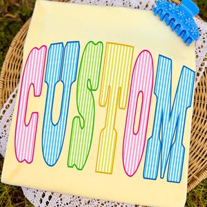 May include: A pale yellow t-shirt featuring the word CUSTOM printed in large, colorful, vertically striped letters. The letters alternate between pink, blue, green, and yellow. A bright blue plastic hair claw clip with a floral design is attached.