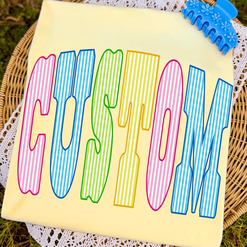 May include: A pale yellow t-shirt featuring the word CUSTOM printed in large, colorful, vertically striped letters. The letters alternate between pink, blue, green, and yellow. A bright blue plastic hair claw clip with a floral design is attached.