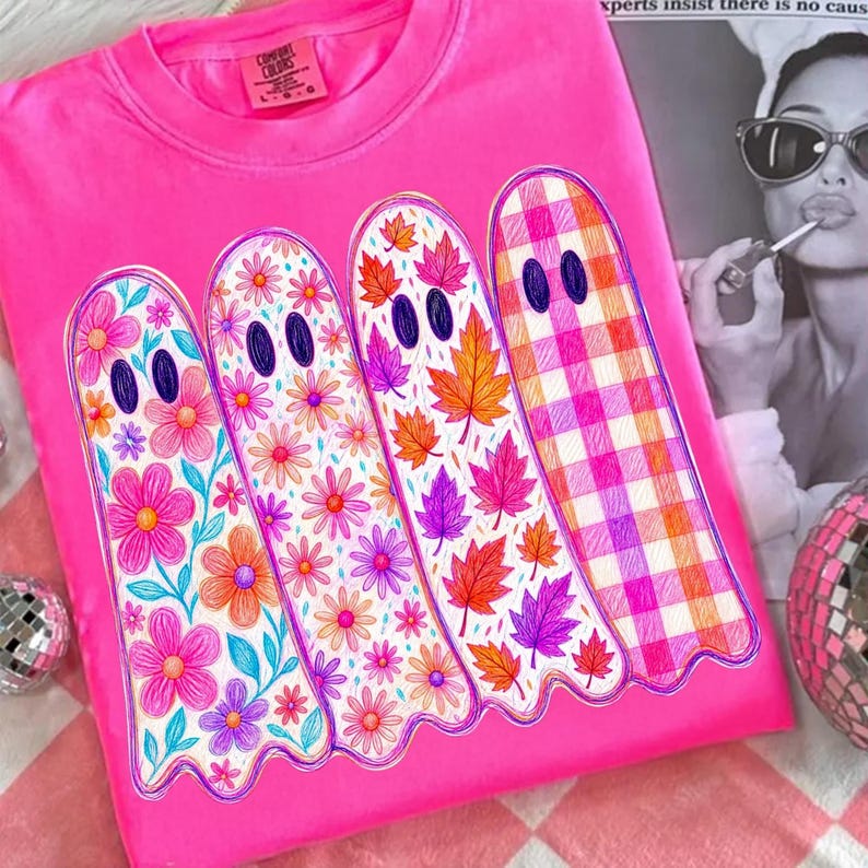 May include: A bright pink t-shirt featuring a graphic of four ghosts standing side by side. Each ghost has a unique pattern: one with pink and orange flowers, one with smaller flowers, one with autumn leaves, and one with a pink and orange gingham check.