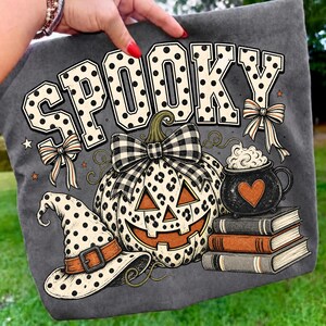 May include: A dark gray t-shirt featuring the word SPOOKY in large polka-dot letters. Below, a leopard-print pumpkin with a black and white plaid bow sits next to a witch hat, a stack of three books, and a black cauldron with a heart and foam.