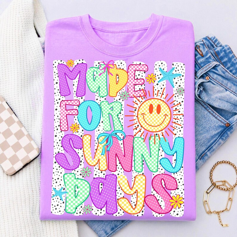 May include: A light purple t-shirt featuring the text "MADE FOR SUNNY DAYS" in colorful, patterned bubble letters. The design includes a yellow smiley face sun, small flowers, and blue starfish icons over a background of vertical black dotted stripes.