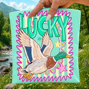 May include: A light mint green tote bag featuring a graphic of a flying mallard duck with brown, white, and green feathers. The word LUCKY is printed in large teal letters above the duck, surrounded by pink squiggly borders and yellow stars.
