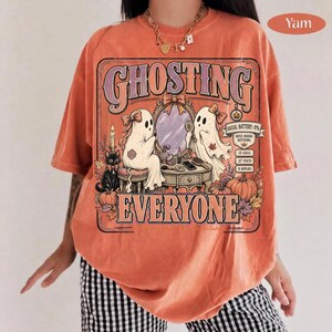 May include: An orange t-shirt featuring a graphic print with the text GHOSTING EVERYONE. The design includes two ghosts, a black cat, a vanity mirror, pumpkins, and a social battery meter reading 0% with 12 calls, 27 texts, and 0 replies.