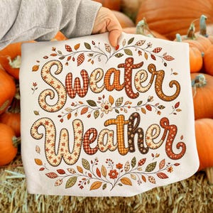 Pu&ograve; includere: Una t-shirt bianca con la scritta Sweater Weather in un grande carattere corsivo decorativo. Le lettere sono riempite con vari motivi a tema autunnale, tra cui quadri, fiori e pois in tonalit&agrave; arancione, marrone e crema.