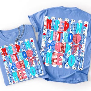 May include: A light blue t-shirt featuring a graphic print with the text "RISED ON DIRT ROADS AND FREEDOM" in red, pink, and blue patterns. The design includes white vertical stripes with black dots, blue bows, and small firework illustrations.