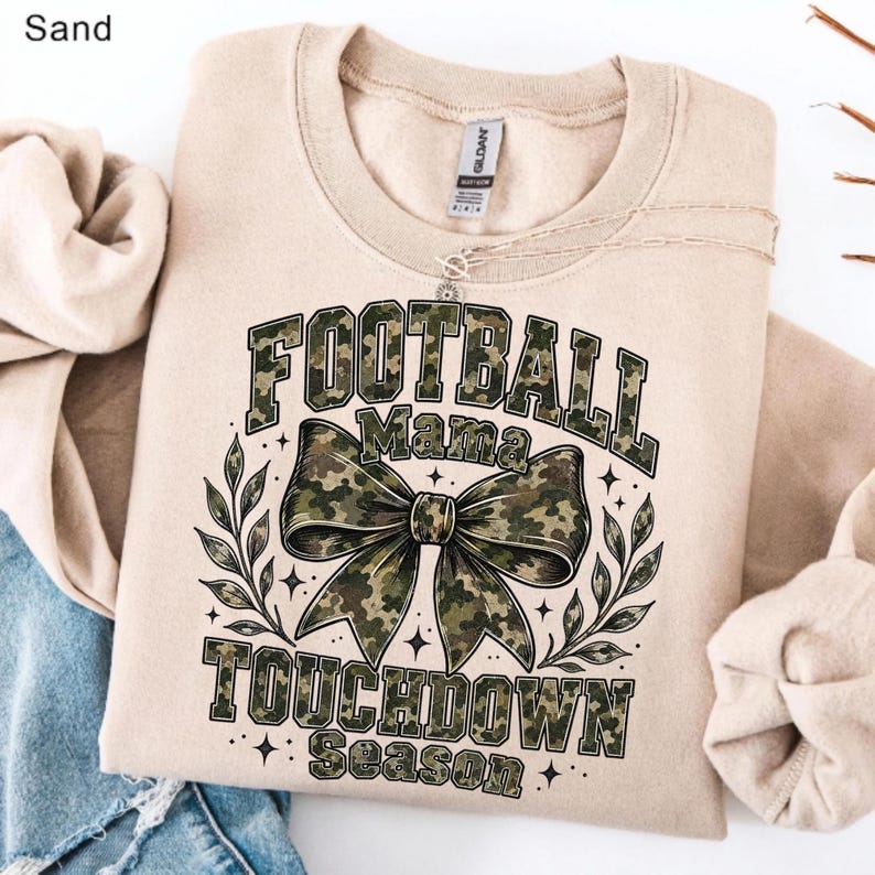 May include: A sand-coloured crewneck sweatshirt featuring a graphic print with the text "FOOTBALL MAMA TOUCHDOWN SEASON" in a camouflage pattern. The design includes a large camouflage bow in the centre, flanked by leafy branches and stars.