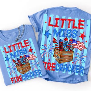 Może przedstawiać: Jasnoniebieski t-shirt z nadrukiem graficznym z napisem "LITTLE MISS FIRECRACKER" w kolorze czerwonym i niebieskim. Wz&oacute;r przedstawia drewnianą skrzynkę wypełnioną czerwonymi, białymi i niebieskimi fajerwerkami, małą amerykańską flagę oraz niebieskie gwiazdki.