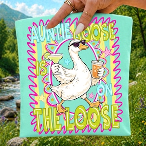 K&ouml;nnte beinhalten: Eine t&uuml;rkisgr&uuml;ne Tragetasche mit einer Cartoon-Gans, die eine Sonnenbrille tr&auml;gt und eine Margarita und ein Getr&auml;nk mit einem Strohhalm h&auml;lt. Der Text "Auntie Goose is on the Loose" ist in einer farbenfrohen, verspielten Schriftart dargestellt.