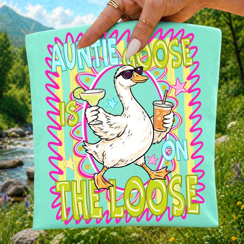 K&ouml;nnte beinhalten: Eine t&uuml;rkisgr&uuml;ne Tragetasche mit einer Cartoon-Gans, die eine Sonnenbrille tr&auml;gt und eine Margarita und ein Getr&auml;nk mit einem Strohhalm h&auml;lt. Der Text "Auntie Goose is on the Loose" ist in einer farbenfrohen, verspielten Schriftart dargestellt.