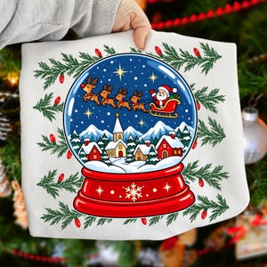 May include: A white t-shirt featuring a graphic of a snow globe with a red base. Inside the globe, Santa Claus in a sleigh pulled by four reindeer flies over a snowy village with a church and houses. Green pine branches with red lights surround the globe.