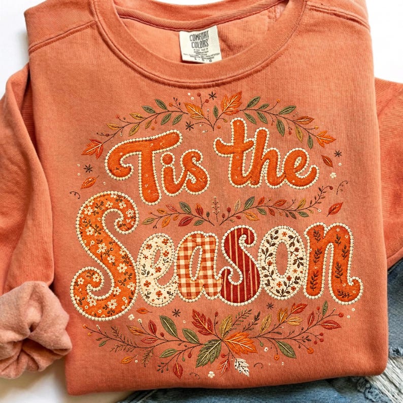 May include: A terracotta-colored crewneck sweatshirt featuring the text 'Tis the Season' in a large, decorative font. The letters are filled with various patterns including floral, gingham, and stripes, surrounded by an embroidered autumn leaf wreath.