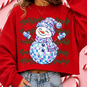 May include: A red long-sleeve sweatshirt featuring a graphic of a snowman made of blue, purple, and white disco ball tiles. The snowman wears a matching hat, scarf, and mittens, set against a background of green pine branches on the red fabric.