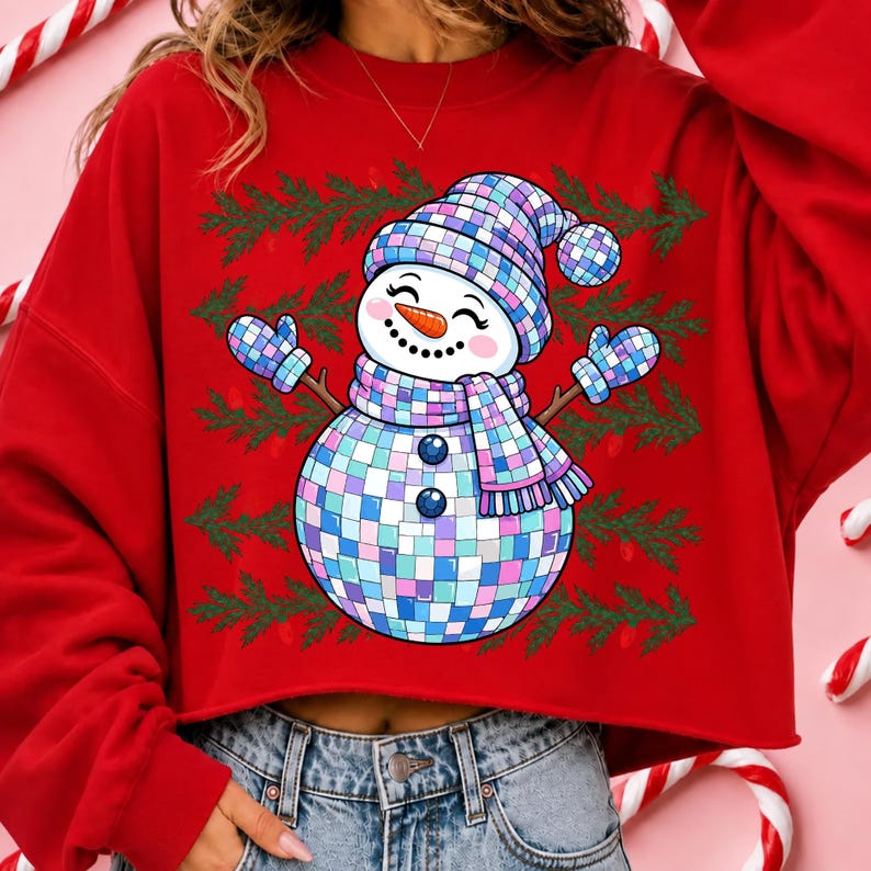 May include: A red long-sleeve sweatshirt featuring a graphic of a snowman made of blue, purple, and white disco ball tiles. The snowman wears a matching hat, scarf, and mittens, set against a background of green pine branches on the red fabric.
