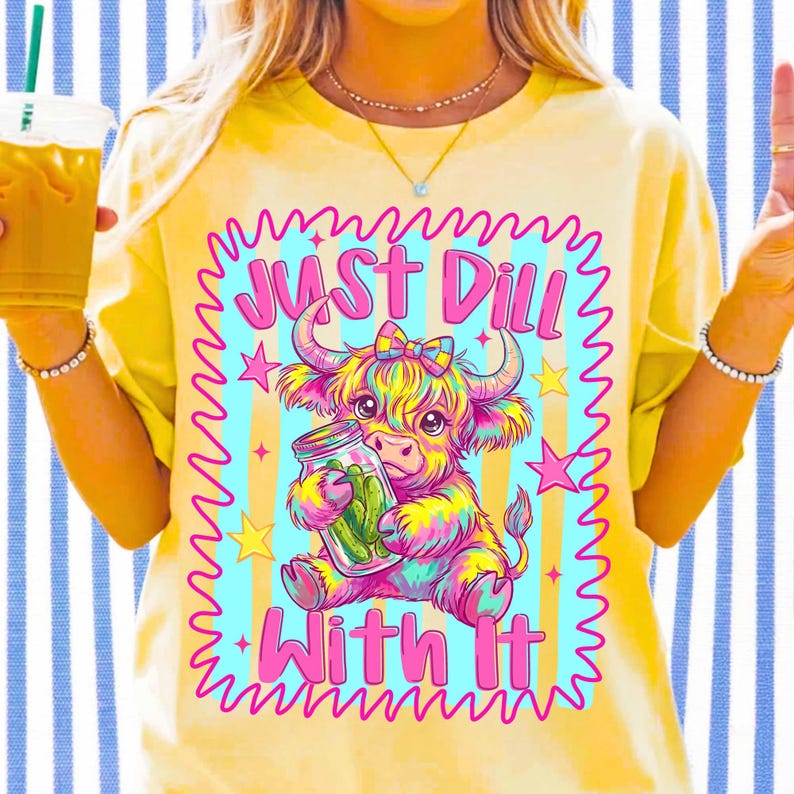 Puede incluir: Una camiseta amarilla con un gr&aacute;fico colorido de una vaca de las Highlands sosteniendo un frasco de pepinillos. El texto "JUST DILL WITH IT" est&aacute; impreso en rosa sobre y debajo de la vaca. El dise&ntilde;o est&aacute; enmarcado por un borde rosa ondulado.