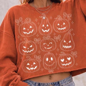 May include: A burnt orange cropped sweatshirt featuring a white line art print of nine various jack-o'-lanterns with different facial expressions, interspersed with small stars and bows. The garment has a crew neckline and long sleeves.