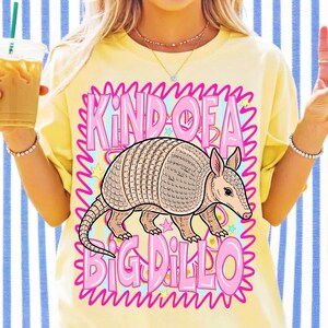Pu&ograve; includere: Una t-shirt giallo pallido con la grafica di un armadillo centrata all'interno di una cornice rosa dai bordi ondulati. Il testo "KIND OF A BIG DILLO" &egrave; stampato in grandi lettere rosa in grassetto sopra e sotto l'illustrazione.