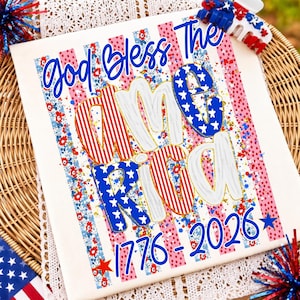 K&ouml;nnte beinhalten: Ein wei&szlig;es T-Shirt mit einem patriotischen Aufdruck mit dem Text God Bless The America 1776-2026 in blauer Schrift. Das Design zeigt vertikale Streifen mit roten, wei&szlig;en und blauen Blumenmustern sowie Sternmotiven.