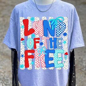 May include: A light gray short-sleeved t-shirt featuring a graphic print with the text "LAND OF THE FREE" in a checkered pattern of red, white, and blue. The design includes small blue stars, light blue flowers, and red and blue firework icons.