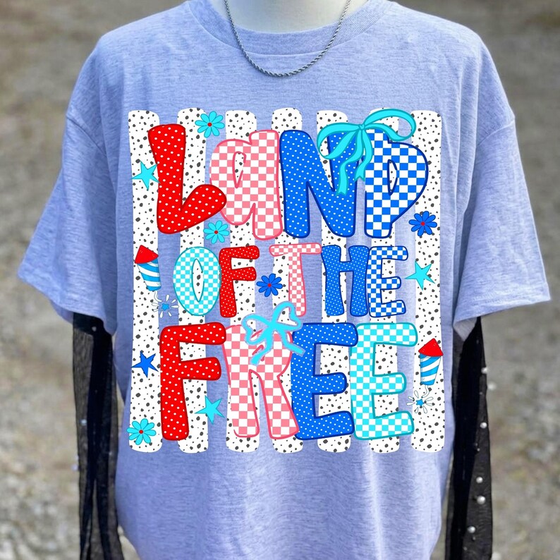 May include: A light gray short-sleeved t-shirt featuring a graphic print with the text "LAND OF THE FREE" in a checkered pattern of red, white, and blue. The design includes small blue stars, light blue flowers, and red and blue firework icons.