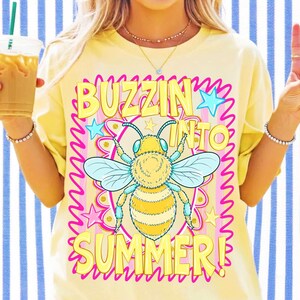 Peut inclure: Un t-shirt jaune p&acirc;le avec un motif central repr&eacute;sentant une abeille jaune et bleue. Le texte BUZZIN INTO SUMMER est &eacute;crit en lettres capitales jaunes. Le design comprend une bordure festonn&eacute;e rose, des &eacute;toiles bleues et jaunes.