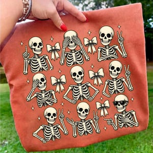 May include: A burnt orange tote bag featuring a repeating pattern of white skeleton figures in various poses, including some wearing sunglasses, making peace signs, or covering their eyes, interspersed with small white bows and four-pointed stars.