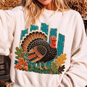 May include: A cream-colored crewneck sweatshirt featuring a large, detailed embroidered graphic of a turkey. The turkey has black, orange, and yellow feathers, a red wattle, and stands amidst green grass, colorful autumn oak leaves, and two acorns.