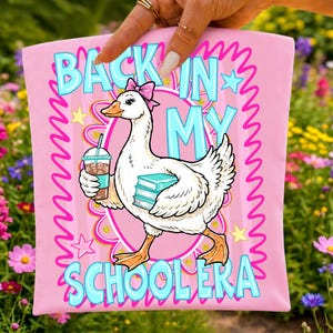 May include: Pink t-shirt with a cartoon goose wearing a pink bow, carrying books and a drink. The shirt has the text "BACK IN MY SCHOOLERA" in blue and white, with star and zig-zag accents.